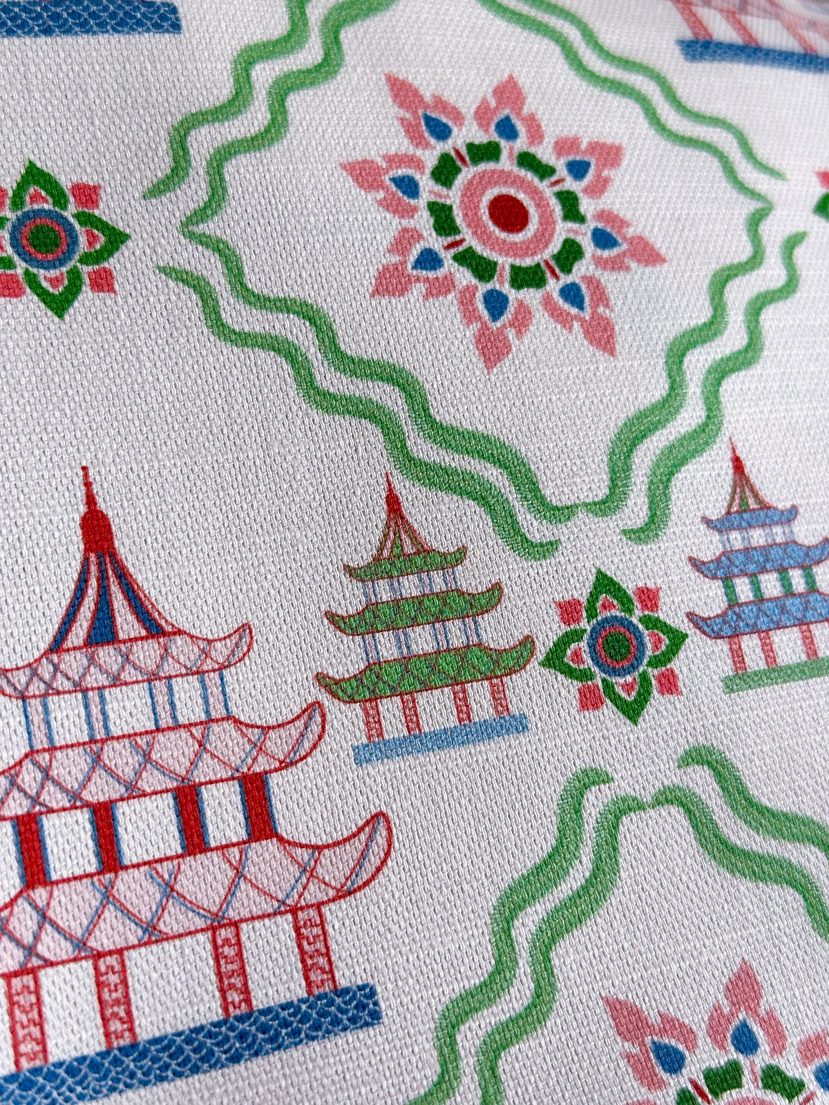A close-up of a white woven fabric printed with a repeating pattern of colorful pagodas, green wavy lines, and floral motifs.